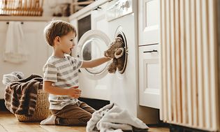 Pojke lägger in nallebjörn i en tvättmaskin i en tvättstuga med tvätt på golvet