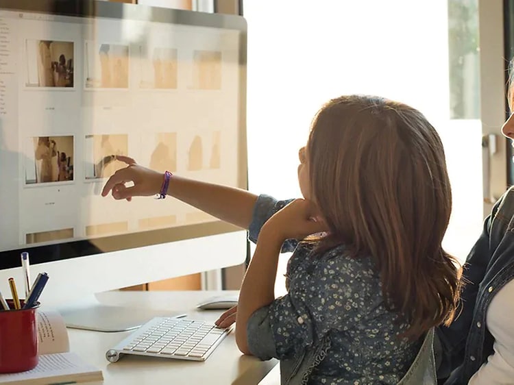 En liten tjej sitter i en kvinnas knä vid ett skrivbord och pekar på bilder på en datorskärm. 