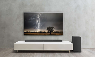 TV med soundbar och basenhet. Åskoväder på tv skärmen, vit tv-bänk och grå vägg bakom.