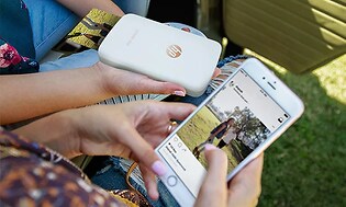 Två personer sitter i en park och håller i en HP Sprocket printer och en vit mobiltelefon med fotoapp öppen. 