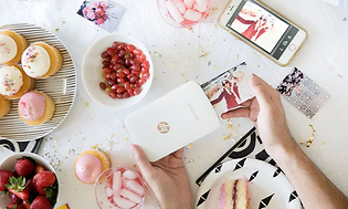 HP Sprocket printer på ett vitt bord med cupcakes och en vit mobiltelefon. En bild är på väg ut från fotoskrivaren.