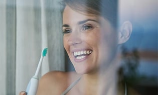 En kvinna står med en eltandborste i handen och ser ut genom fönstret, hon ler stort. 