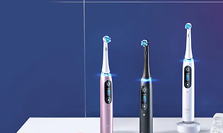 Oral-B iO eltandborstar i tre olika färger. En rosa, en svart ch en vit som står på ett vitt bord med blå bakgrund.
