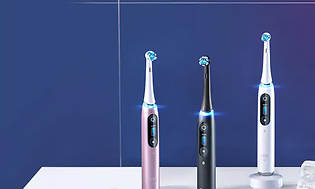 Oral-B iO eltandborstar i tre olika färger. En rosa, en svart ch en vit som står på ett vitt bord med blå bakgrund. 