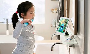 En liten flicka borstar tänderna med en eltandborste i ett badrum samtidigt som hon ser på barnprogram på en surfplatta. 
