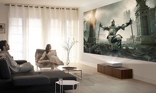 Vardagsrum med två personer som sitter och tittar på film som visas på väggen med hjälp av en projektor. 