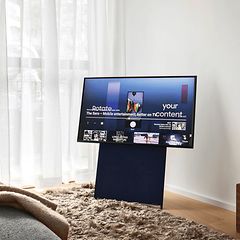 Samsung The Sero på stativ i vardagsrum med stora fönster, ljusa gardiner och en grå soffa. 