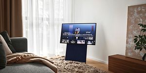 Samsung The Sero på stativ i vardagsrum med stora fönster, ljusa gardiner och en grå soffa. 