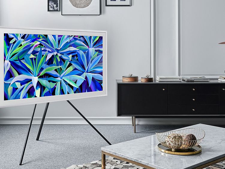 En vit Samsung TV, The Frame 2021, på stativ i hörnet av ett vardagsrum. Ett bord står framför och en svart bänk i bakgrunden.