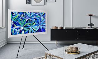 En vit Samsung TV, The Frame 2021, på stativ i hörnet av ett vardagsrum. Ett bord står framför och en svart bänk i bakgrunden.