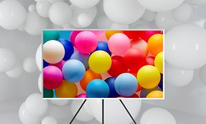 Samsung TV The Frame 2021 på stativ med färgglada ballonger på skärmen och vita ballonger i bakgrunden. 