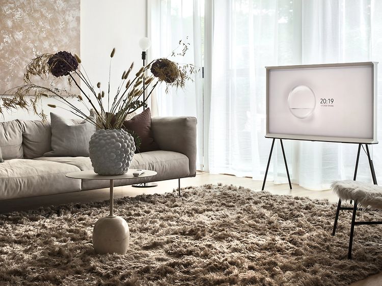Samsung The Serif TV på stativ i ett ljust vardagsrum med stora fönster och ljusinsläpp genom tunna gardiner. Beiga möbler.