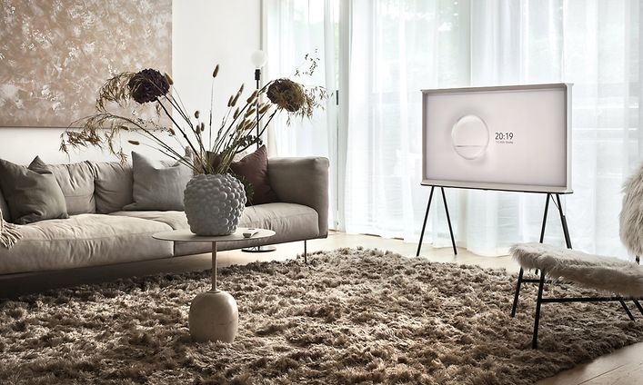 Samsung The Serif TV på stativ i ett ljust vardagsrum med stora fönster och ljusinsläpp genom tunna gardiner. Beiga möbler.  