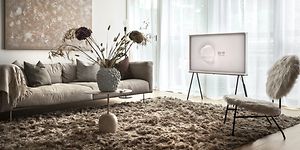 Samsung The Serif TV på stativ i ett ljust vardagsrum med stora fönster och ljusinsläpp genom tunna gardiner. Beiga möbler.  