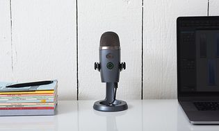 En mikrofon av modellen Blue Yeti Nano på ett vitt skrivbord med en hög böcker till vänster och en laptop till höger.