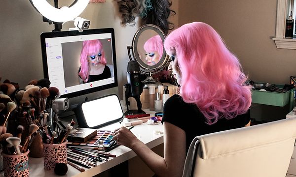 Kvinna med rosa hår sminkar sig vid ett sminkbord med en dator framför sig med en filmkamera och en mikrofon vid sidan. 