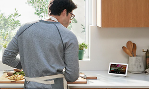 Ryggtavlan på en man som står i ett kök och lagar mat med hjälp av Google Nest Hub på skärmen.