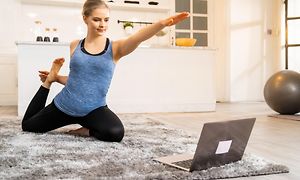 Kvinna gör yoga på en matta på golvet med en bärbar dator uppfälld framför dig, i bakgrunden syns en pilatesboll. 