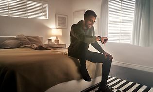 En man sitter på kanten på en stor säng och kollar på klockan på armen, en svart Garmin Venu. 