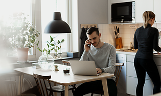 En man som arbetar på sin bärbara dator och pratar i sin smartphone i ett kök