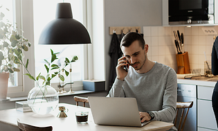 En man som arbetar på sin bärbara dator och pratar i sin smartphone i ett kök