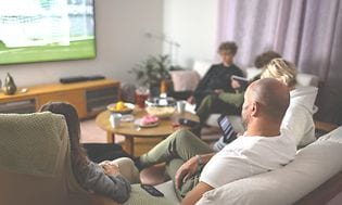 Familj i soffa som tittar på sport på TV och har snacks på ett runt bord i mitten
