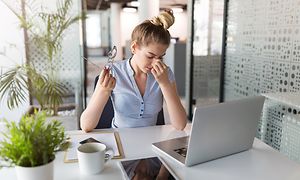 Kvinna vid sin laptop som håller sig mot näsan och blundar och visar att hon inte mår bra