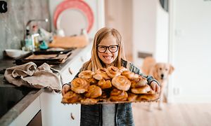 Glad flicka håller en bricka full med kanelbullar i ett kök och en hund i bakgrunden