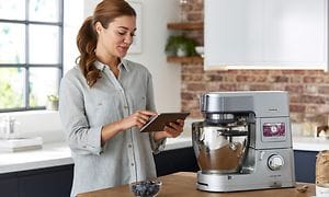 En kvinna står med en surfplatta och använder Kenwood World-appen bredvid en Kenwood Cooking Chef XL köksmaskin