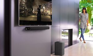 kvinna går bredvid en golvhögtalare i en gång med en TV-skärm på väggen med en soundbar under