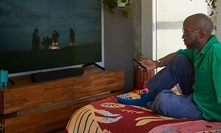 Man som sitter på en säng i ett sovrum med en fjärrkontroll i handen och tittar på TV
