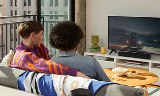 Sonos-Par som tittar på TV i en soffa med höga hus utanför fönstret och en högtalare bakom dem