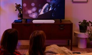 Sonos-Par som tittar på TV i ett mörkt vardagsrum med en vit Sonos soundbar på TV-bänken under TV:n