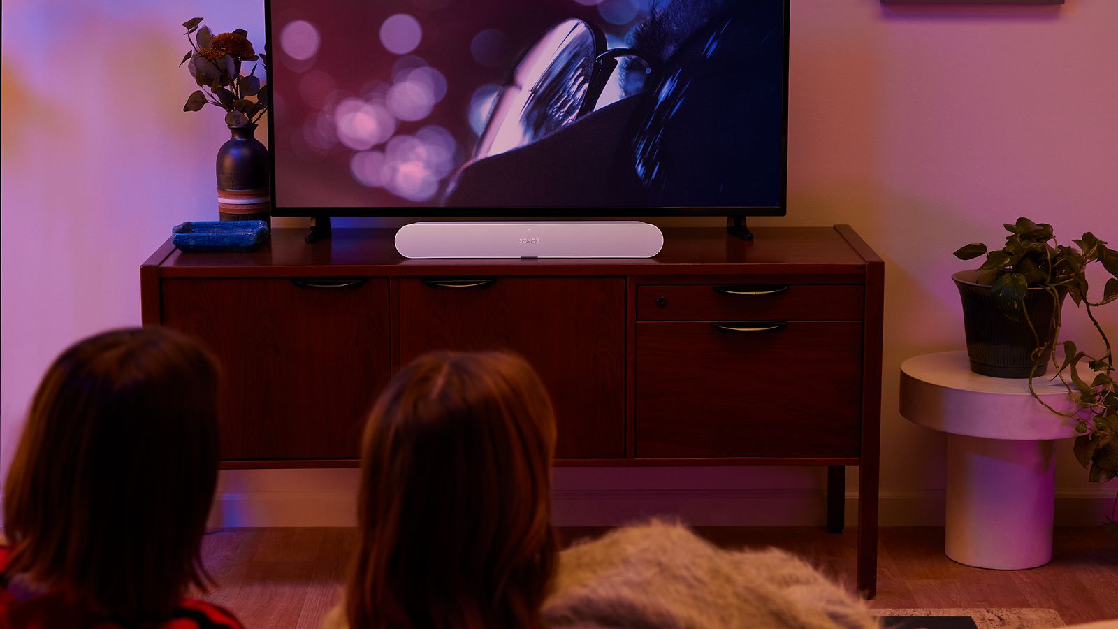 Sonos-Par som tittar på TV i ett mörkt vardagsrum med en vit Sonos soundbar på TV-bänken under TV:n