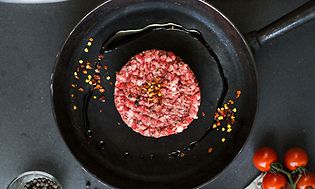 Hamburgerkött på en stekpanna sett uppifrån och färskt kött och hamburgerbröd på sidan