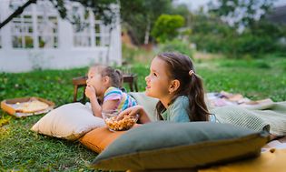 Två barn ligger på kuddar på en gräsmatta med snacks och pizza framför sig och ser ut att titta på TV
