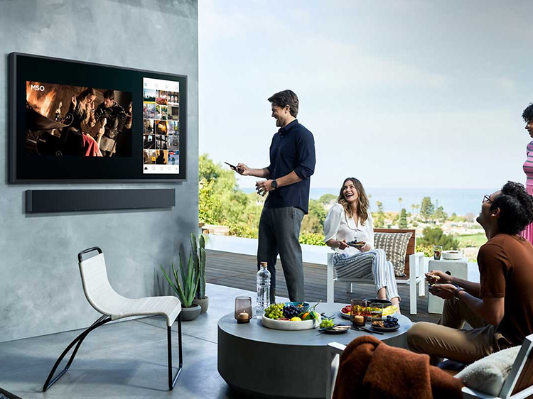 Samsung-Fyra personer tittar på The Terrace TV utomhus på en terrass med frukt på ett bord och med sol och utsikt över landskap