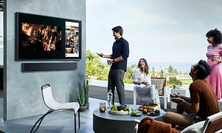 Samsung-Fyra personer tittar på The Terrace TV utomhus på en terrass med frukt på ett bord och med sol och utsikt över landskap
