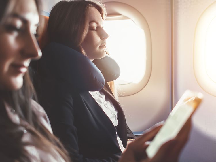 Two women relax on a plane, one with a neck pillow and the other looking at her phone