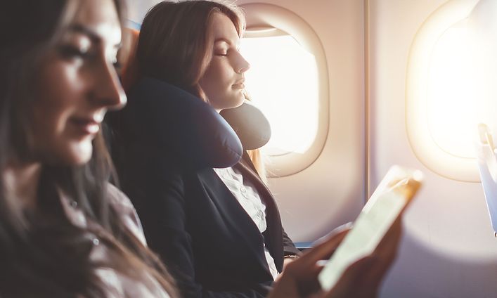 Two women relax on a plane, one with a neck pillow and the other looking at her phone