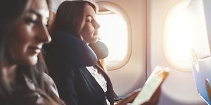 Two women relax on a plane, one with a neck pillow and the other looking at her phone
