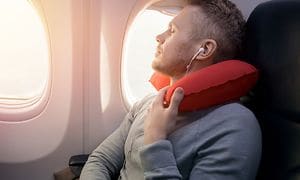 A man is sitting on an aeroplane next to a window sleeping with a neck pillow
