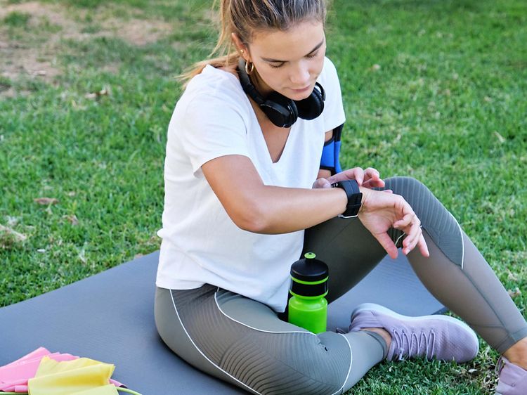En kvinna som sitter på en yogamatta utomhus med vattenflaska och träningsredskap bredvid sig kollar sin smartwatch