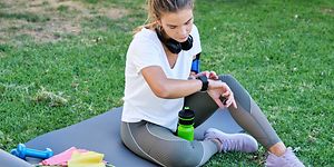 En kvinna som sitter på en yogamatta utomhus med vattenflaska och träningsredskap bredvid sig kollar sin smartwatch