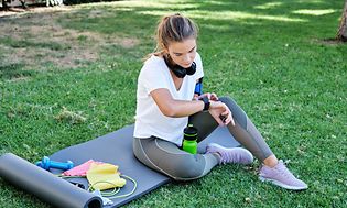 En kvinna som sitter på en yogamatta utomhus med vattenflaska och träningsredskap bredvid sig kollar sin smartwatch