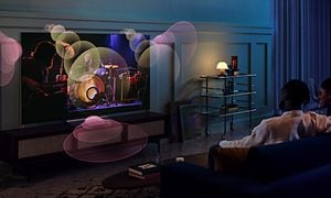 Ett par sitter i en soffa med en TV-skärm framför sig och illustrerade ljudvågor som kommer ur den från olika håll