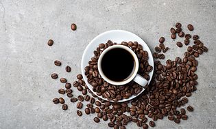 Kaffekopp med kaffebönor utspridda runt om på en grå bordsskiva.