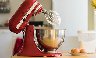 KitchenAid köksmaskin på ett bord, intill ägg och en plastbehållare med socker