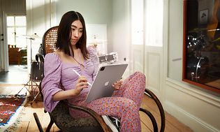 Ung asiatisk kvinna i en gammeldags gungstol och med ett trumset i bakgrunden sitter och jobbar med en iPad Pro med M1-chip