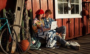 Två tonåringar sitter utomhus vid en röd sjöbod och jobbar vid en MacBook Air